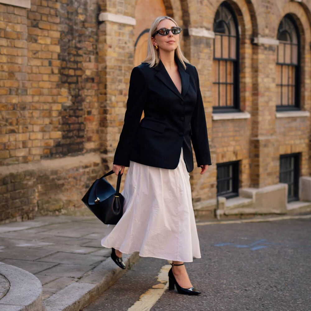 Woman wears white skirt, black blazer, black heels, and a black handbag while posing for photos. 