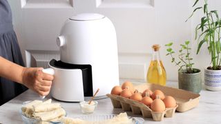 woman holding tray handle of white air fryer on kitchen counter, with eggs on the side