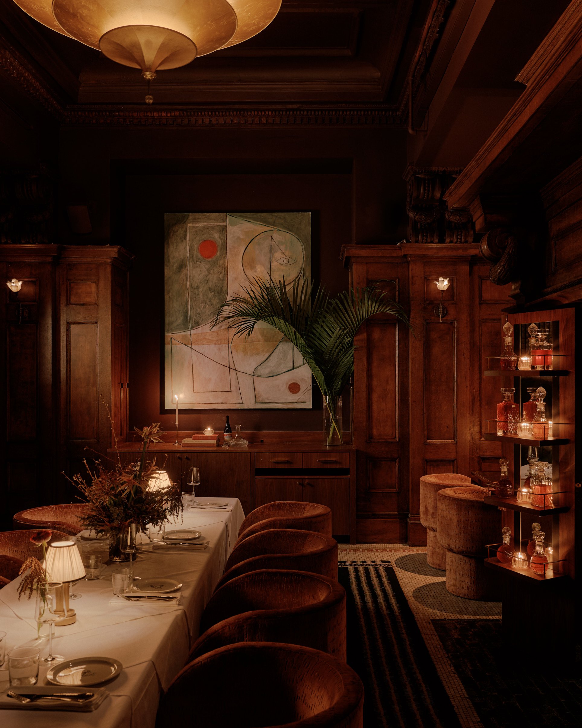 A dimly lit, imaginatively decorated cocktail bar features murals, paintings, Art Deco-inspired lighting, and sprawling plants in a warmly suffused atmosphere.