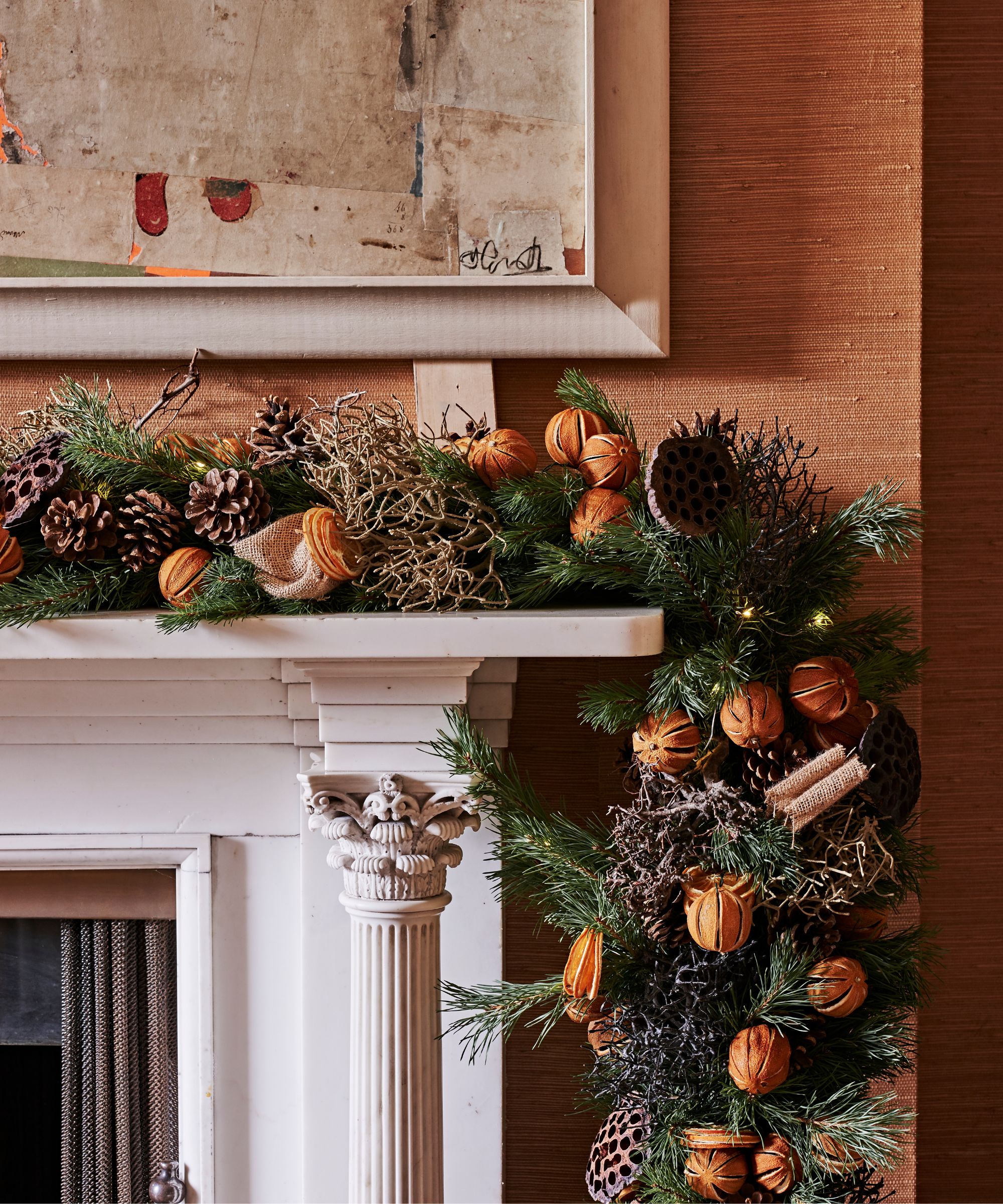 A festive white mantelpiece with pines, oranges and dried pinecones, with orange wallpaper behind and an artwork above.