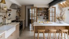 A kitchen with a stone and wood island with wooden stools, a wood and glass pantry, a hanging rod and open shelf for pans and glasses beside the induction hob and oven