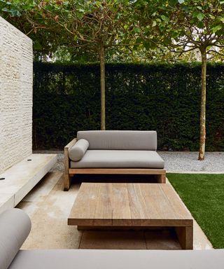 Minimalist outdoor patio furniture ideas with sleek wooden furniture and neat box hedges and trees behind.