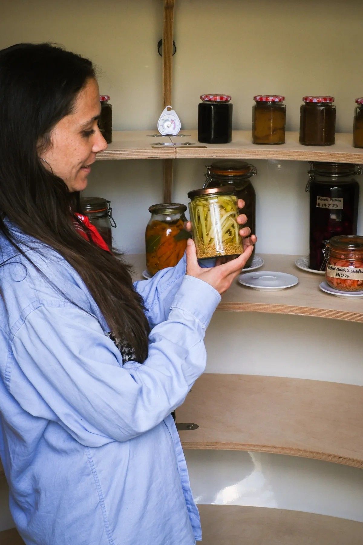 A young woman in a blue shirt and denim jeans opens the lacquered green door to a subterranean fridge in a garden, where a series of pickle jars sits.