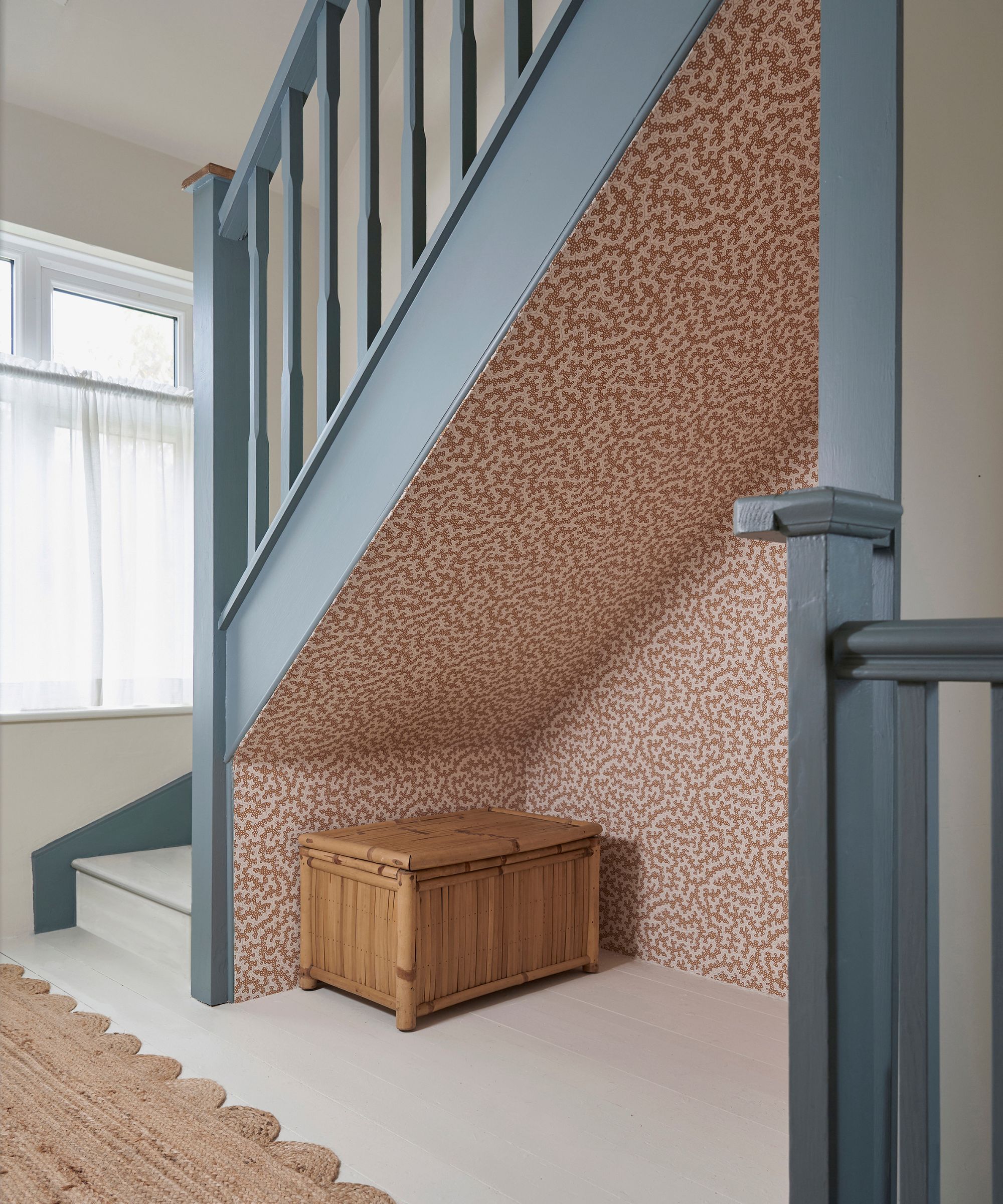 An under-stairs nook with patterned terracotta wallpaper, painted blue banister, and a woven bamboo storage chest on a pale wooden floor.