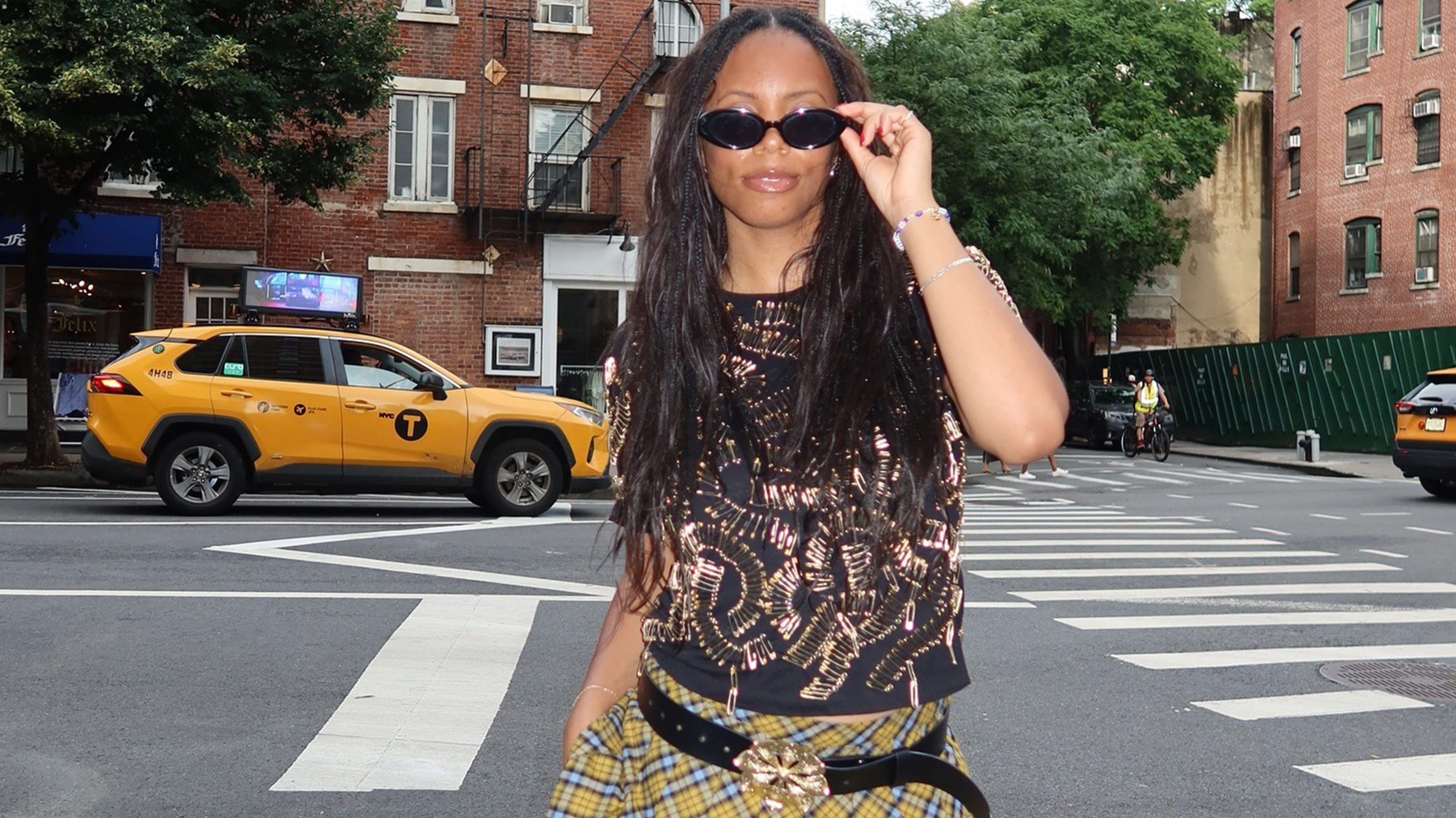 Sierra wears a printed black shirt, black sunglasses, a yellow plaid skirt and black belt with a gold buckle.