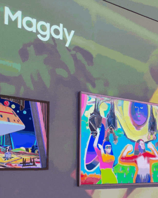A multimedia, audiovisual installation features a background video depicting a hand holding lit up candles, while the wall onto which it is projected showcases a series of striking canvases on digital screens.