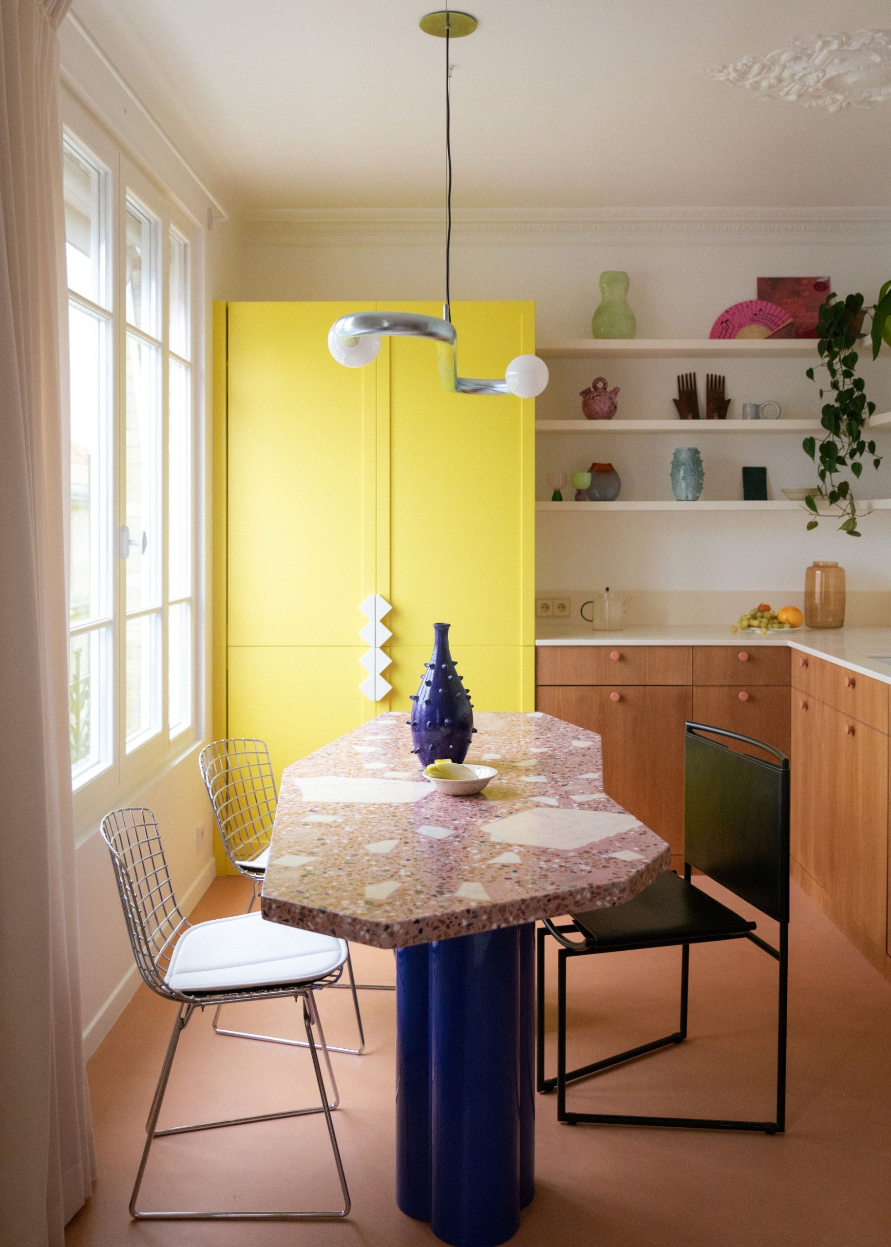 A modern apartment kitchen with white walls, wood cabinets, and a tall dresser painted lemon yellow. Open shelving with decorative items. A terrazzo dining table with an electric blue base and modern metal dining chairs.