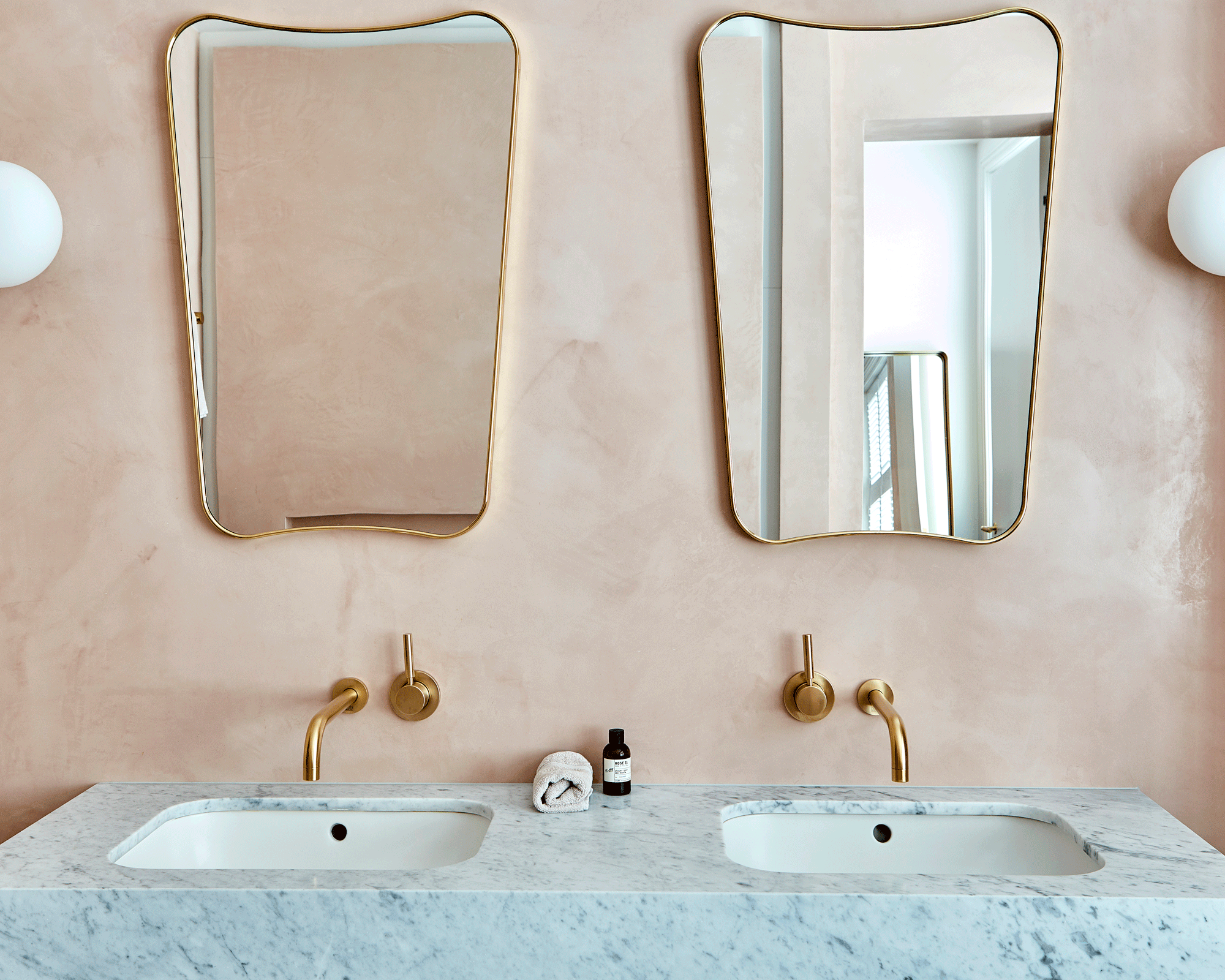 An undermounted double vanity unit with gold faucets and matching mirrors against a pink wall