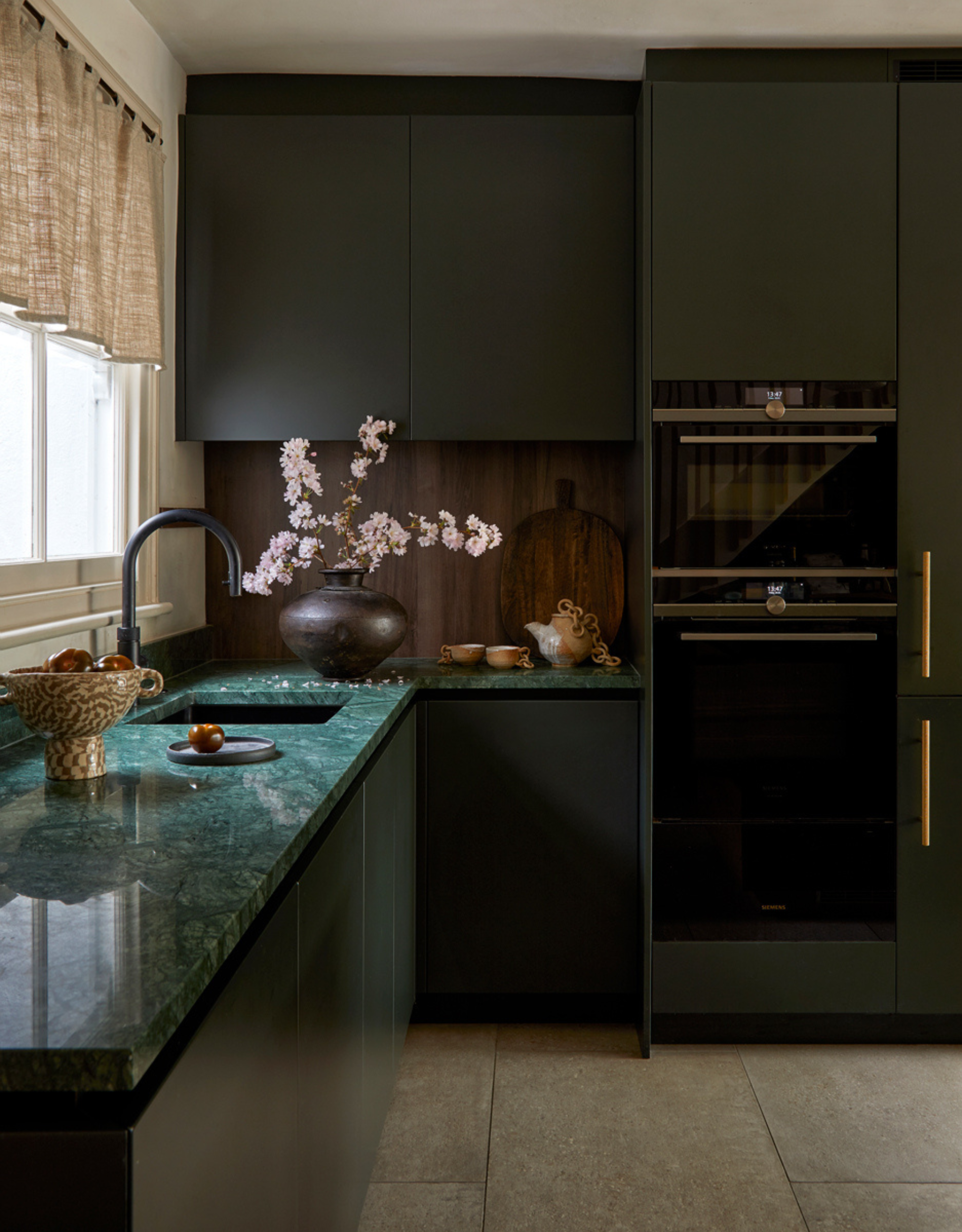A beautiful kitchen with a green marble counter, dark green storage cabinets, a vase of flowers, a bowl of fruit, a wooden backsplash, and a ceramic tea set