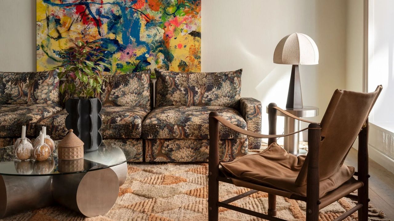 living room with verdure print sofa, orange rug, leather armchair, silver and glass coffee table, chrome side table with lamp, and colorful artwork on wall behind the sofa