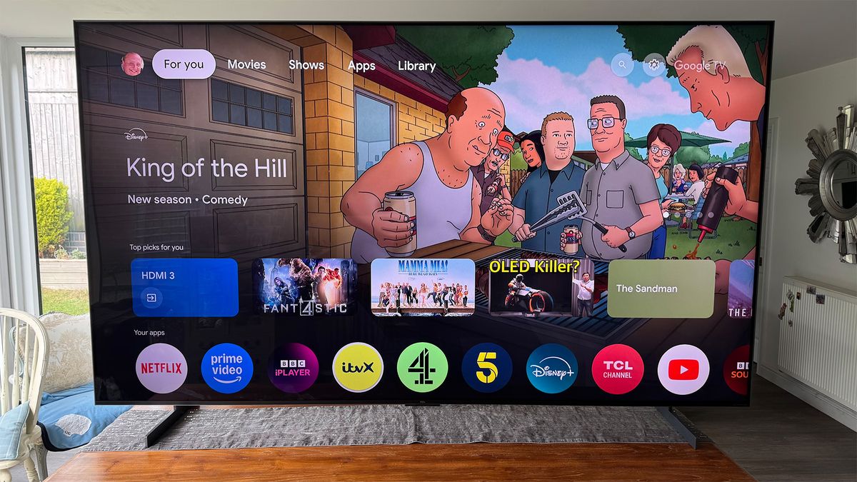 The TCL 98C7K 98-inch TV on a table in a living area, seen straight-on. On screen is a still from King Of The Hill, above a row of other shows/films and a row of apps.