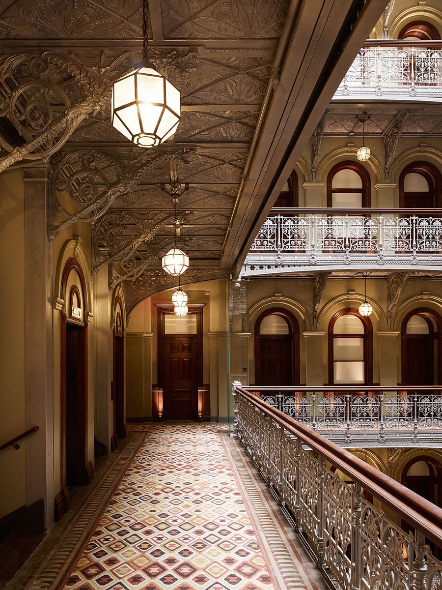 A timeless corridor in a hotel is brought to life by Art Deco lighting, a mosaiced floor, and ornate wood and iron rails.