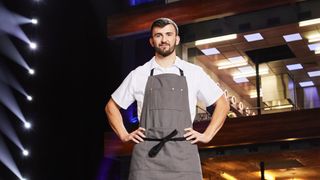Contestant Callum in a white top and grey apron in front of the kitchen in Next Level Chef.
