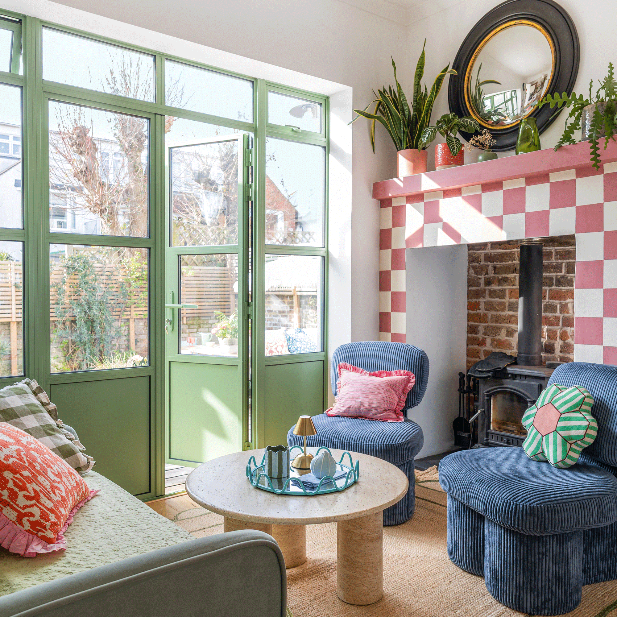 a snug with green patio doors a sofa and two blue corduroy covered chairs with a pink checkered painted fire surround and a logburner