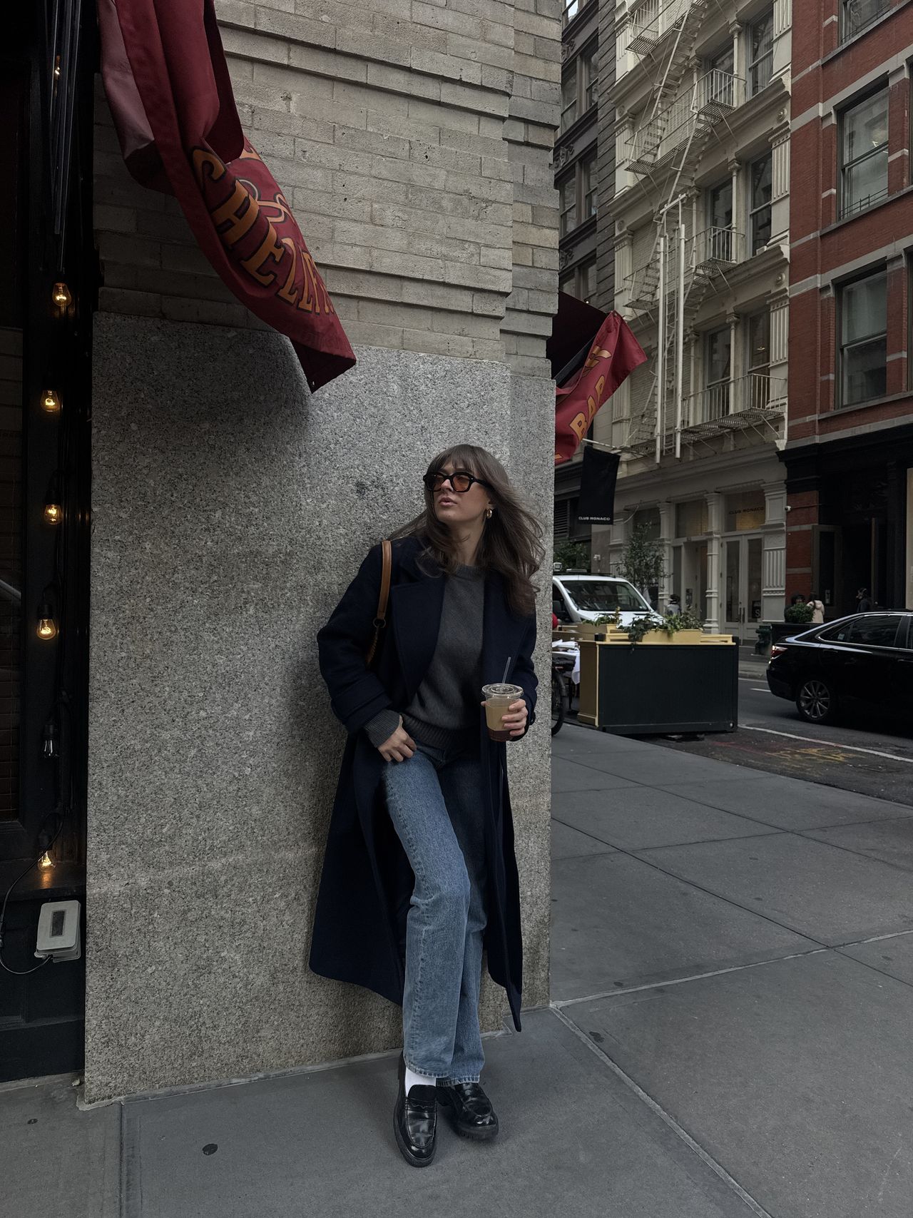 Kathryn Zahorak leaning on wall wearing sunglasses, navy coat, jeans, loafers