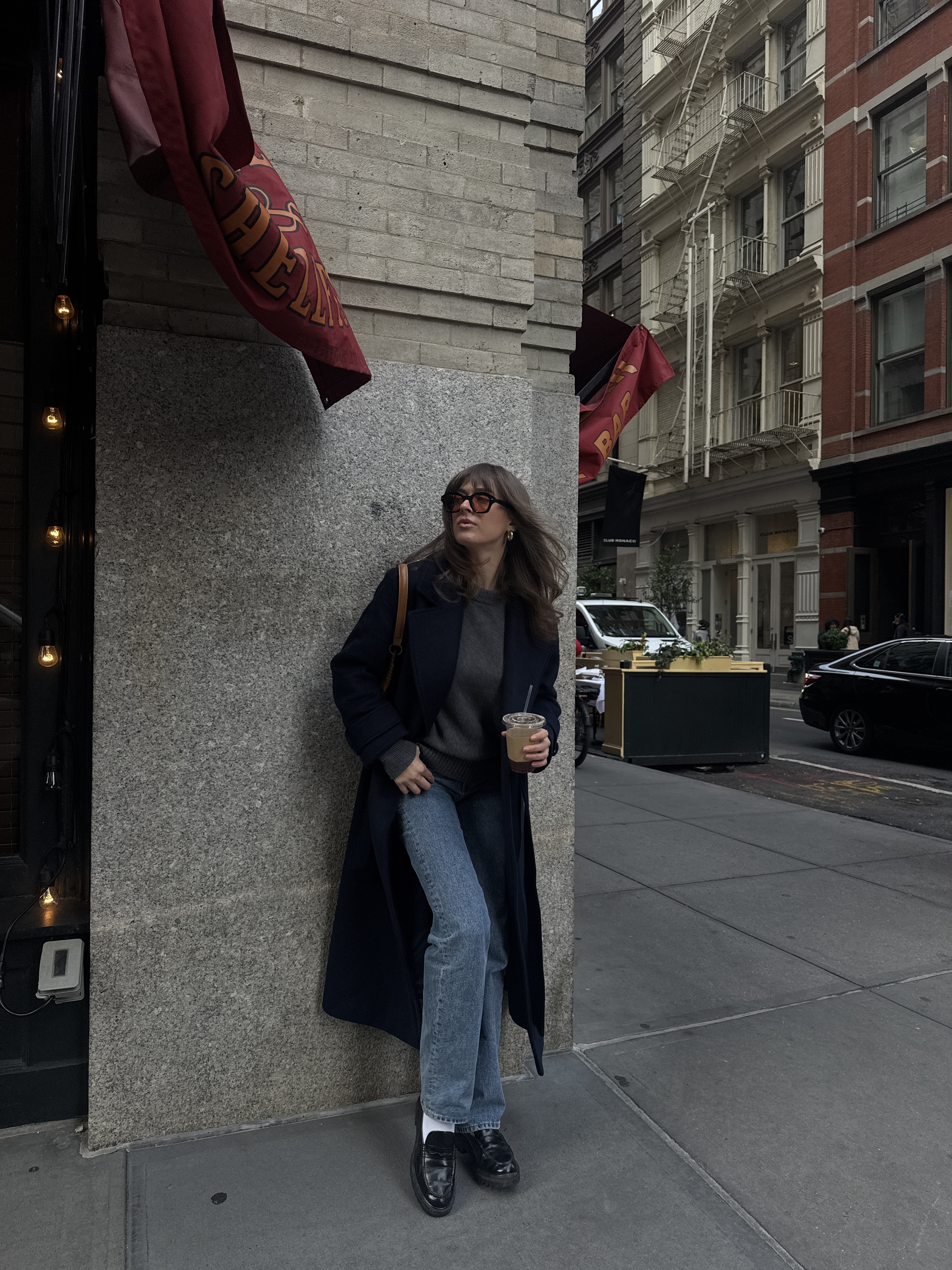 Kathryn Zahorak leaning on wall wearing sunglasses, navy coat, jeans, loafers
