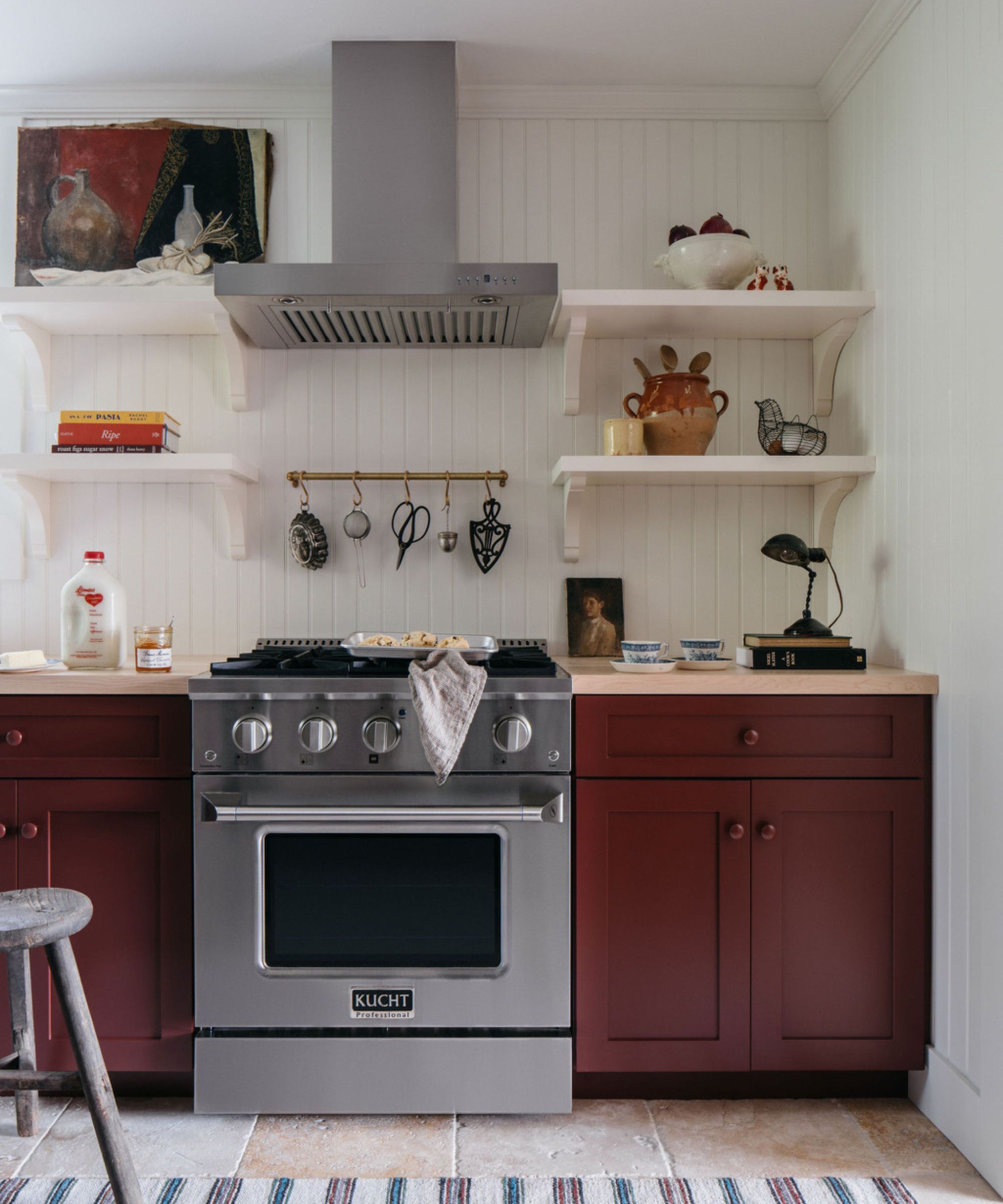 A cottage kitchen with white paneled walls, burgundy cabinets, wooden countertops, stainless steel appliances and white open shelving