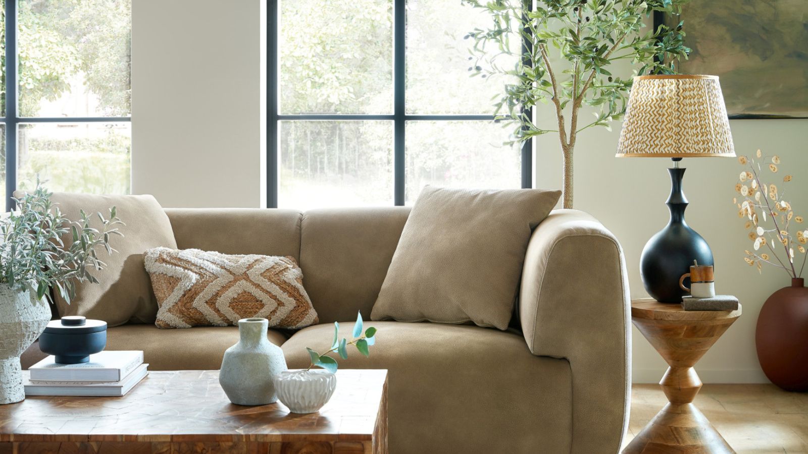 A large mink colored sofa in a white living toom. A small wooden side table with a tall side lamp on top to the right hand side. A wooden coffee table with vases on it in front. 