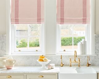 Elegant white kitchen with pink fabric blinds, marble kitchen countertop and splashback, brass fittings