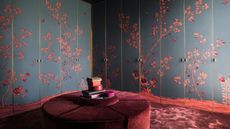 Dark, luxe walk-in closet with floor-to-ceiling built-in wardrobes, matte dark blue wallpapered doors with red oriental floral design. Plush burgundry carpets with round velvet matching large seat in center