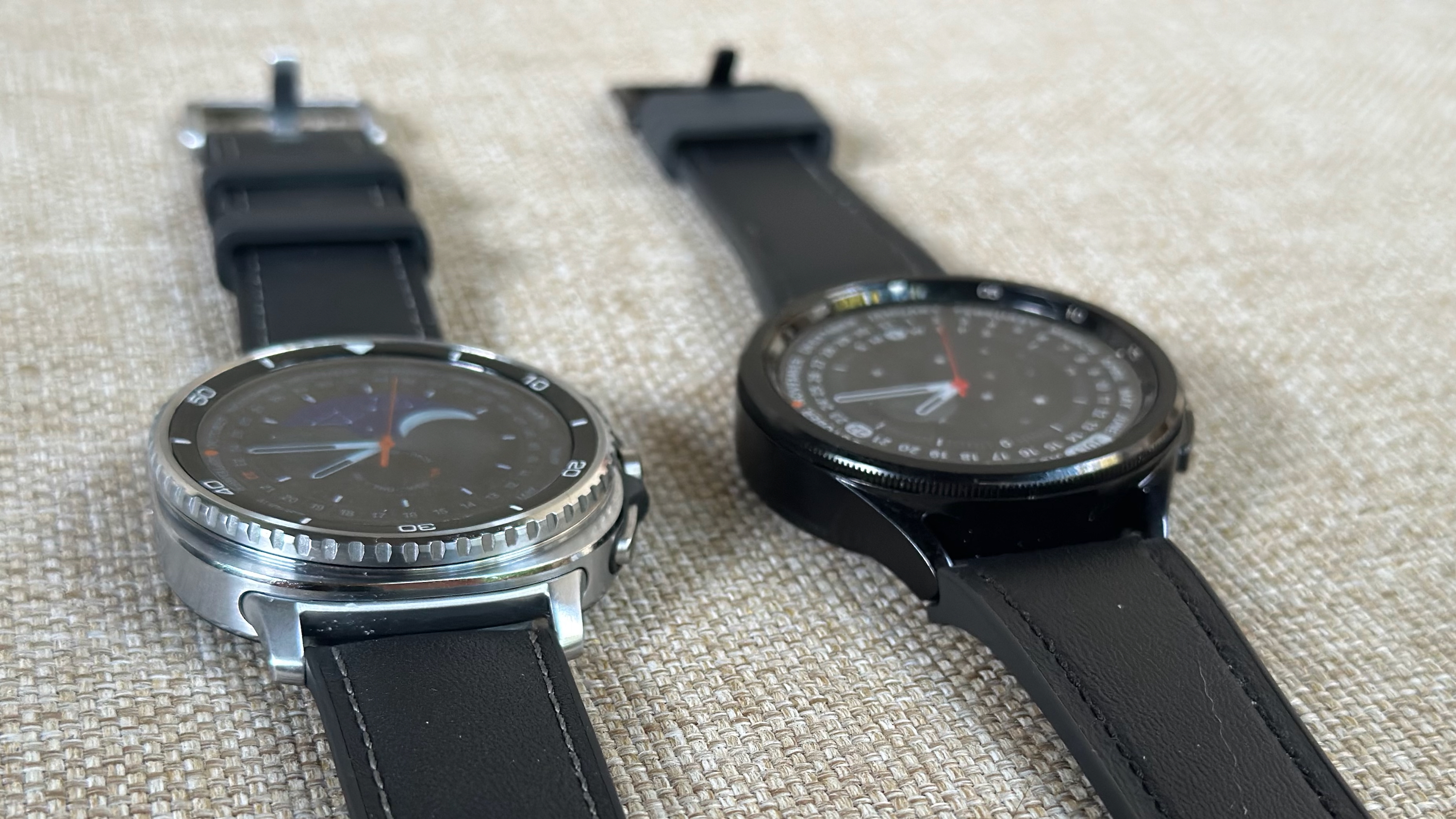 The Samsung Galaxy Watch 8 Classic (left) and Samsung Galaxy Watch 6 Classic (right) sitting aside one another, the photo angled to focus on their rotating bezels and thickness from the bottom-up. 