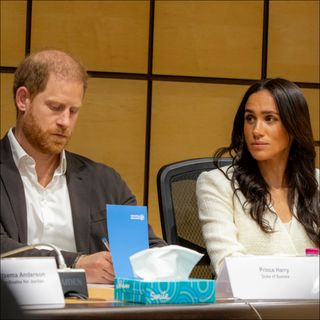Prince Harry wears a suit without a tie and looks somber while wife Meghan Markle wears a white top and has long black hair
