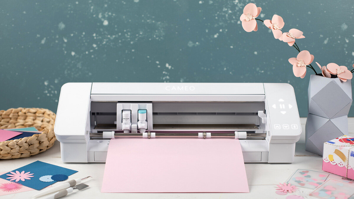 A Silhouette Cameo 4 sits on a desk with paper feeding into it.