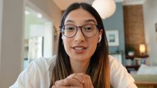 Woman wearing glasses and white earbuds in domestic setting looks at viewer