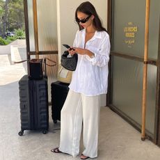 style influencer Felicia Akerstrom poses with suitcases while looking at her phone in front of the Les Roches Rouges hotel in France wearing black sunglasses, an oversize white linen button-down shirt, relaxed ivory silk pants, and a small black woven Bottega Veneta bag