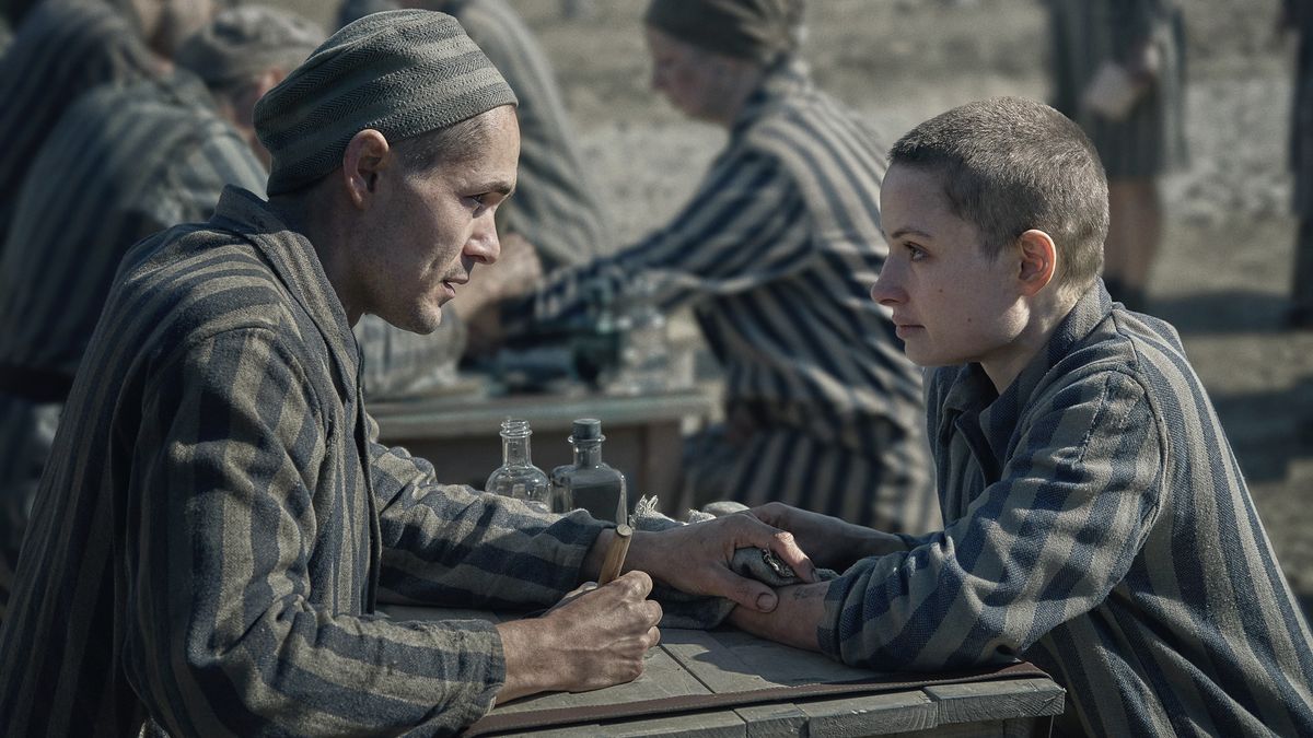 Jonah Hauer-King as Lali sits across a table from Anna Próchniak as Gita, both in striped Auschwitz uniforms in The Tattooist of Auschwitz