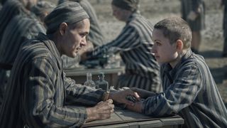 Jonah Hauer-King as Lali sits across a table from Anna Próchniak as Gita, both in striped concentration camp uniforms in The Tattooist of Auschwitz