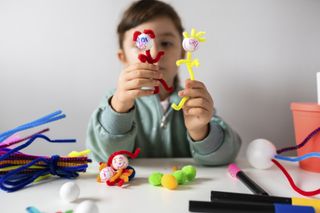 A young playing with two pipe cleaner stick figures and sat at a desk with her crafts.
