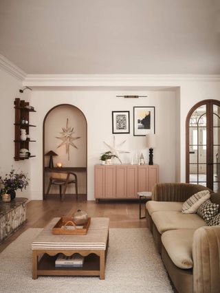 Image of a white living room with pops of pink and sage green. The rug is off-white with a scalloped coffee table/ottoman. There is a velvet, sage green sofa behind the table and dusty pink cabinet on the back wall.