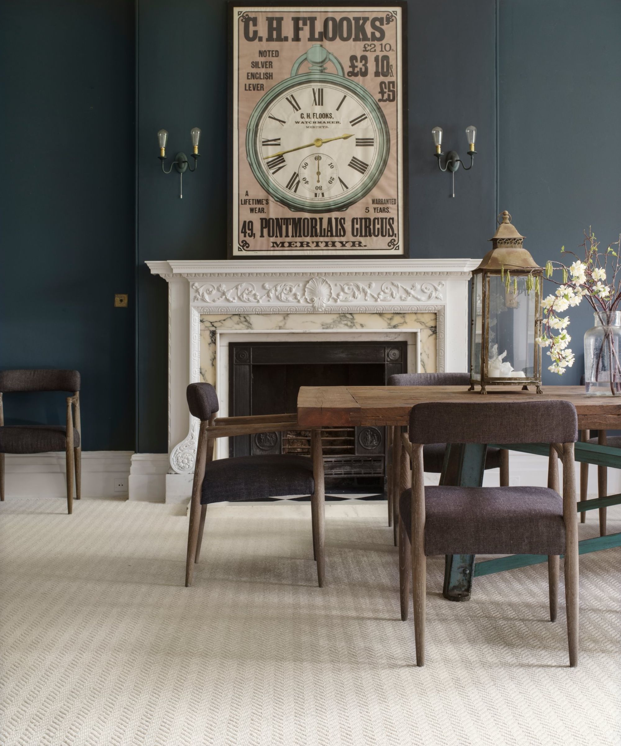 The first image shows a dining room with a deep teal wall and an ornate white fireplace topped by a large vintage-style clock. A rustic wooden dining table with dark upholstered chairs sits on a pale textured carpet, adding contrast and warmth to the elegant space.
