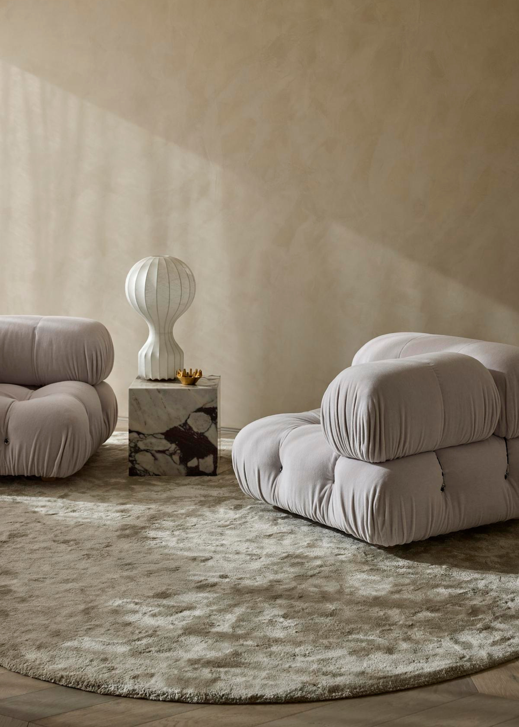 A living room with low upholstered sofa seaters, a travertine side table with a sculptural lamp and a circular plush rug