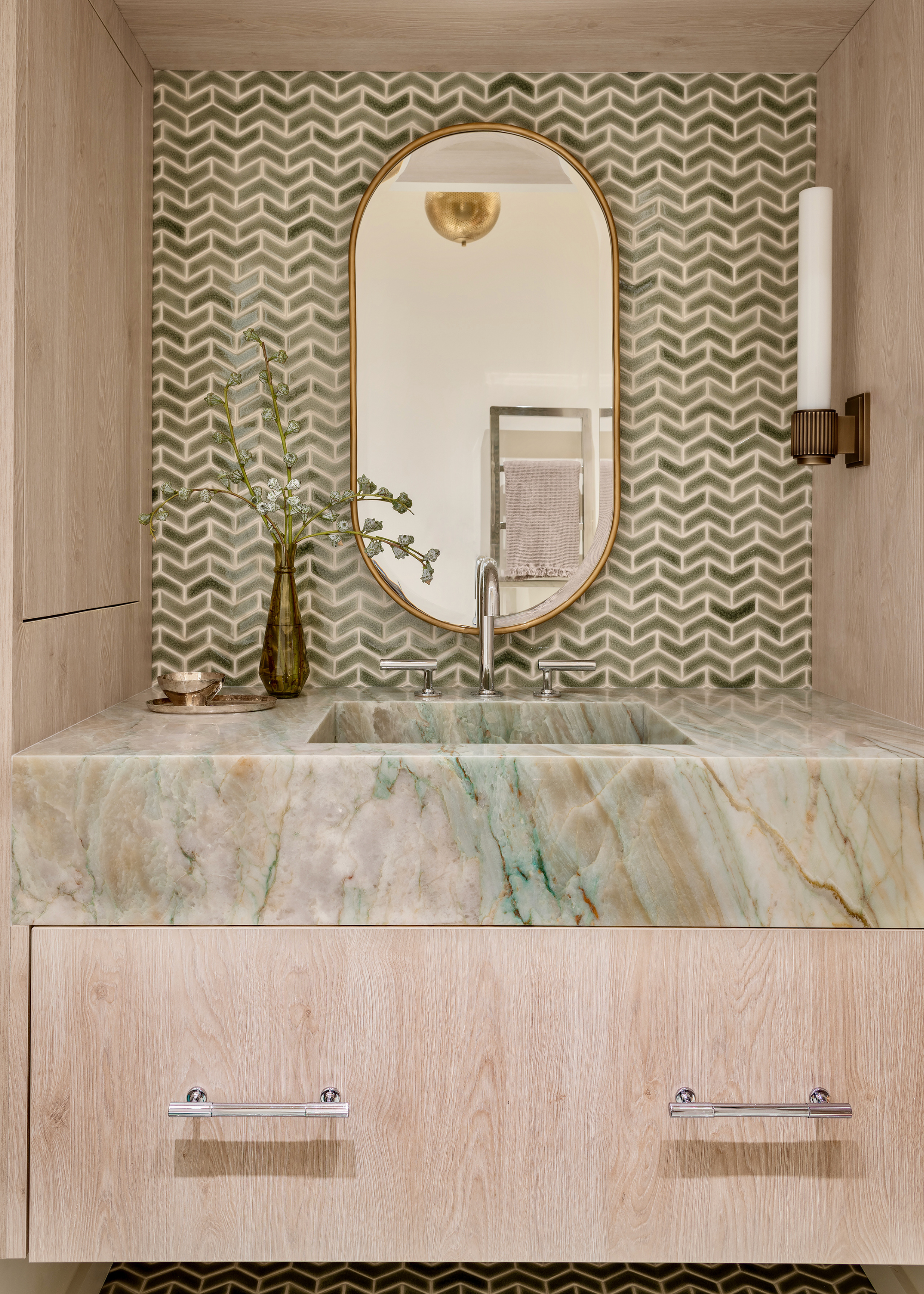 A powder room with mini green tiles in a pattern on the back wall with a brown and green veined marble sink surrounded by pale wood, with a gold-framed bathroom mirror, a wall light and foliage in a vase