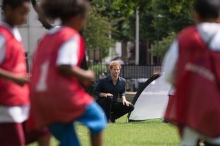 Prince harry kneeling down in grass behind school kids playing soccer