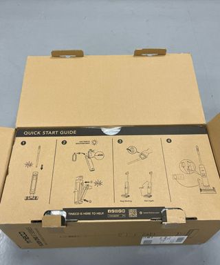 Tineco Floor One S7 Pro cordless wet vacuum's cardboard box, partially open showing quick start guide of how to use the vacuum, on gray floor in Future test center