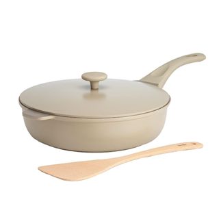 A cream colored frying pan with a lid on top and a wooden spatula in front of it