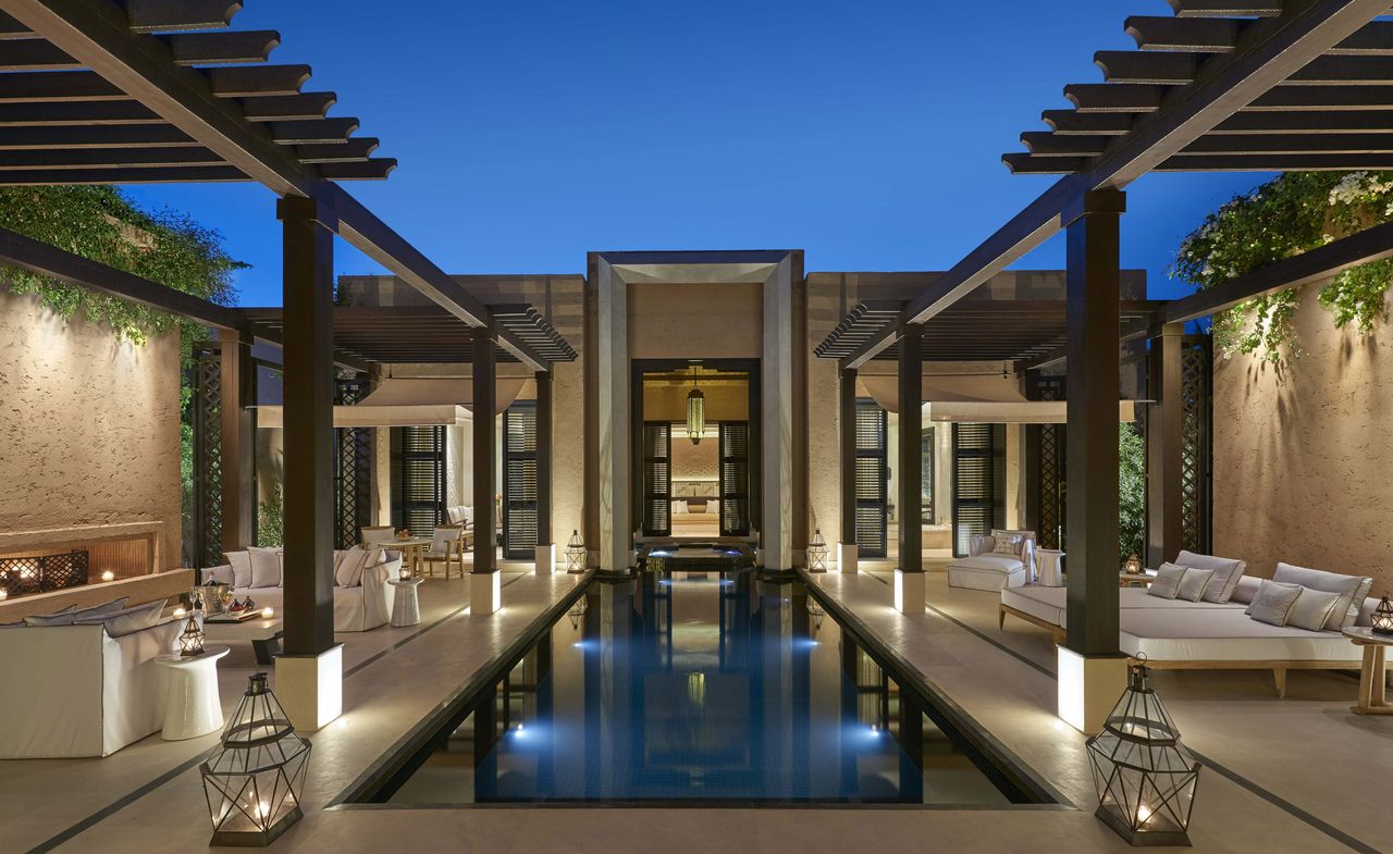 A hotel pool area with, a long pool, sofas, loungers and floor lamps.