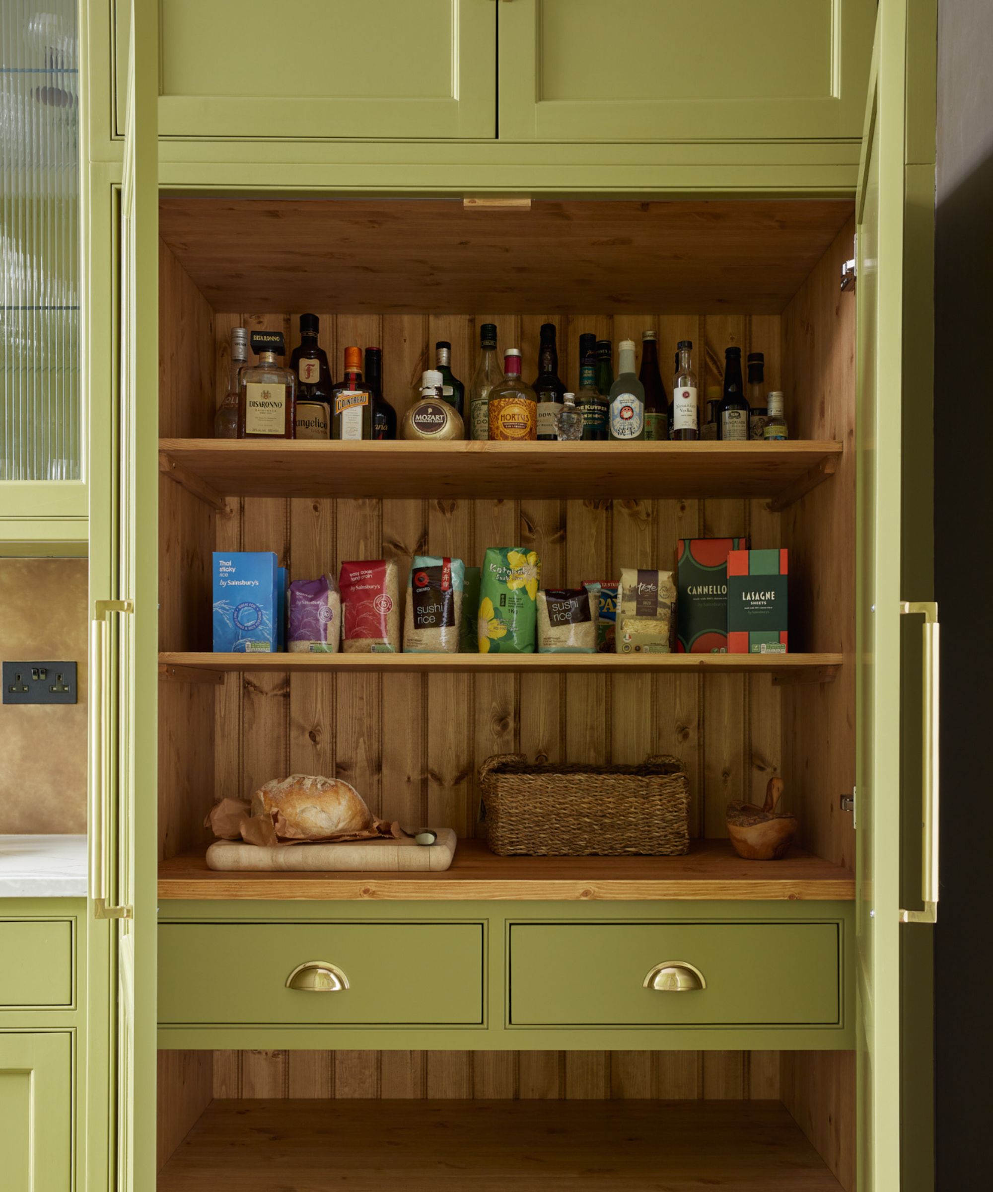 Classic country-style pantry in soft olive green with warm wood interiors, open shelves of dry goods and bottles, and brass cup pulls for a welcoming, traditional look.