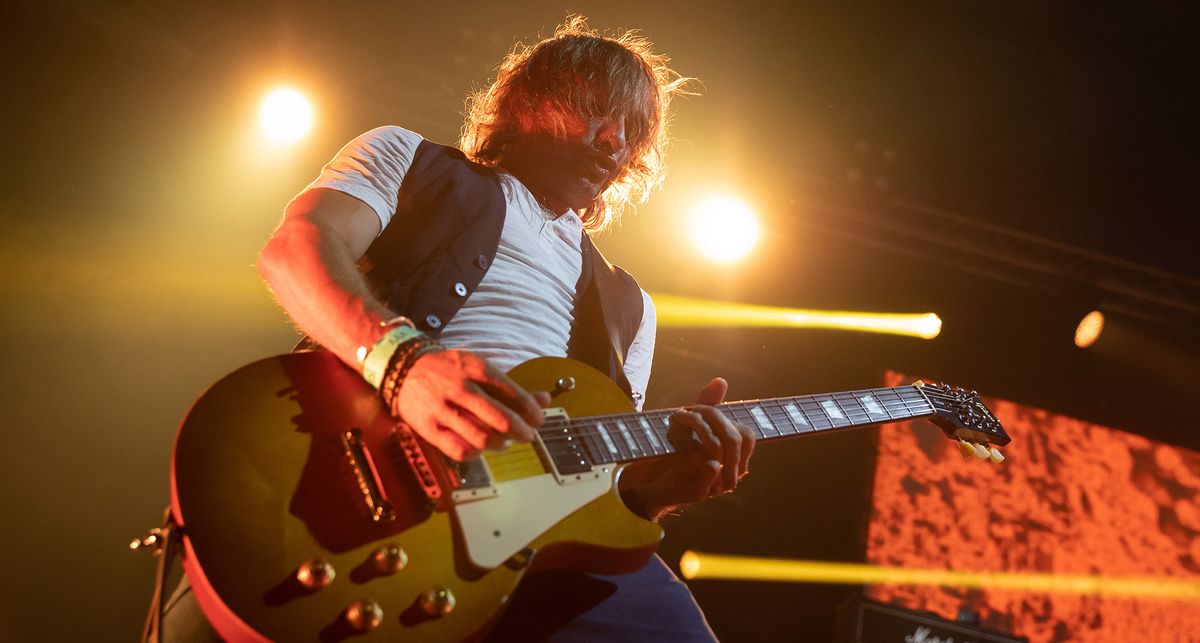 Joe Hoare of Orange Goblin plays a solo his Gibson Les Paul onstage, wearing a white t-shirt and black waistcoat. 