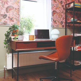 A home office with walls covered in pink floral wallpaper and a dark wood desk paired with a brown leather desk chair