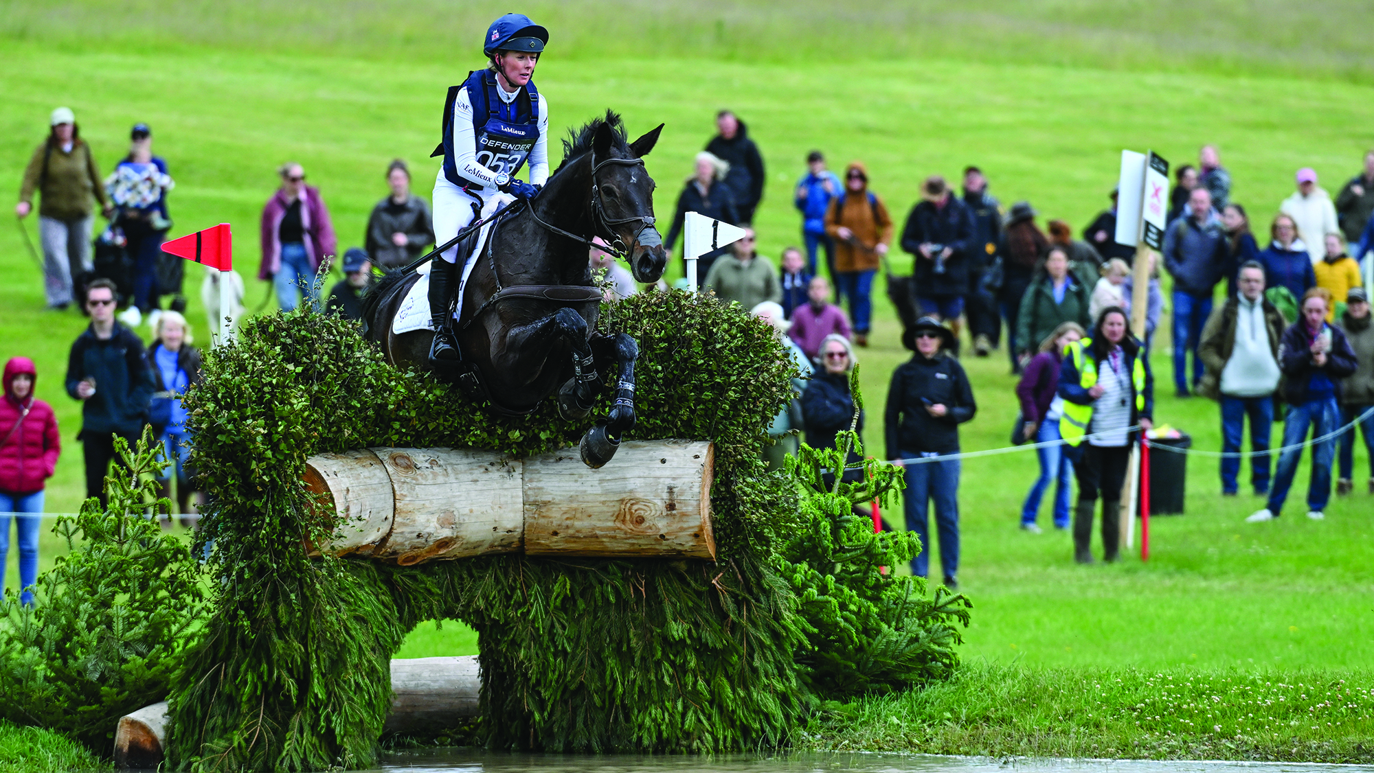 A rider in a blue top with white sleeves and a blue hat rides a dark horse over a brush jump into water at Bramham Horse Trials 2026