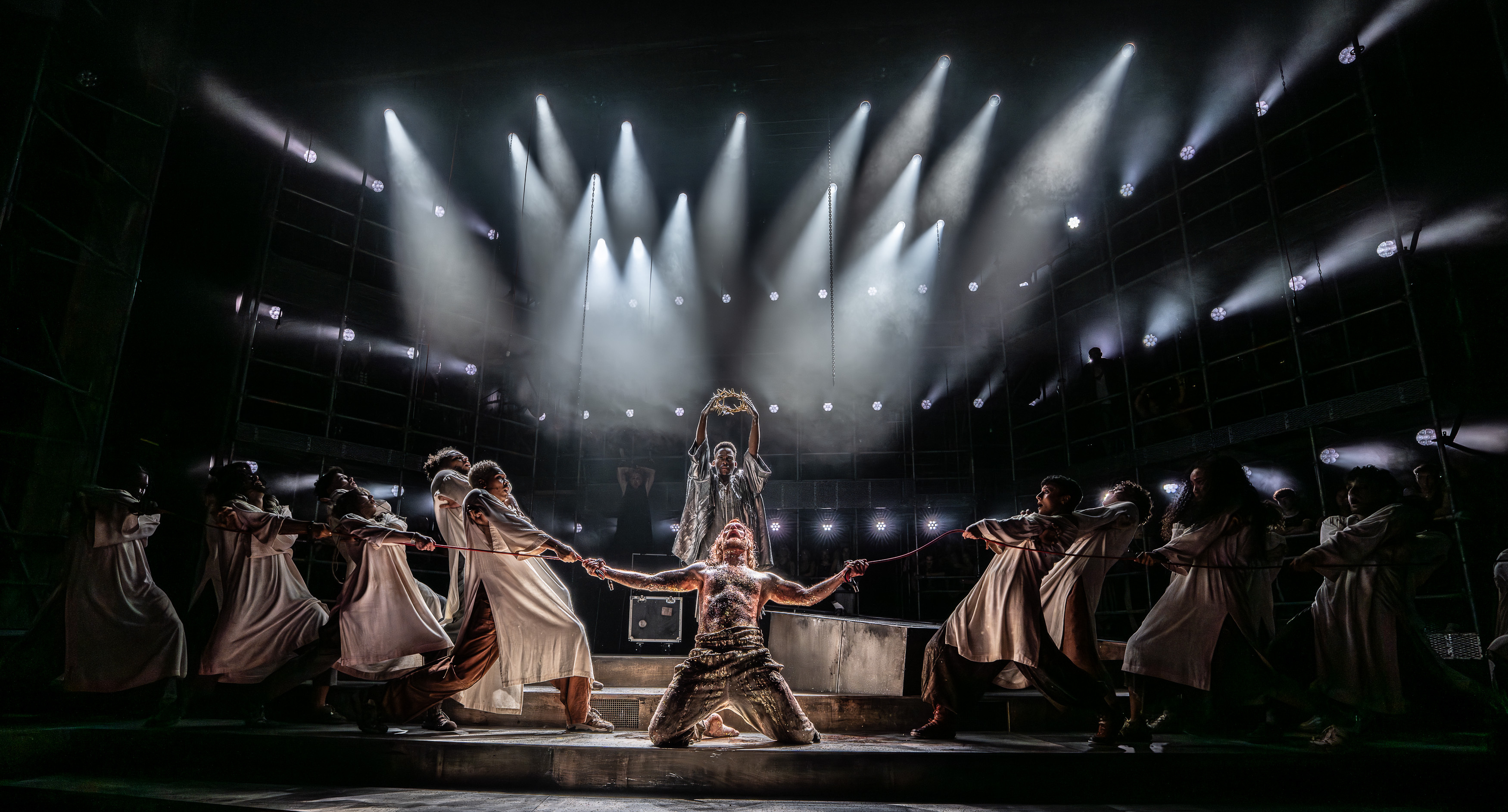 an image of Sam Ryder onstage as Jesus in Jesus Christ Superstar with members of the ensemble being tied to the cross