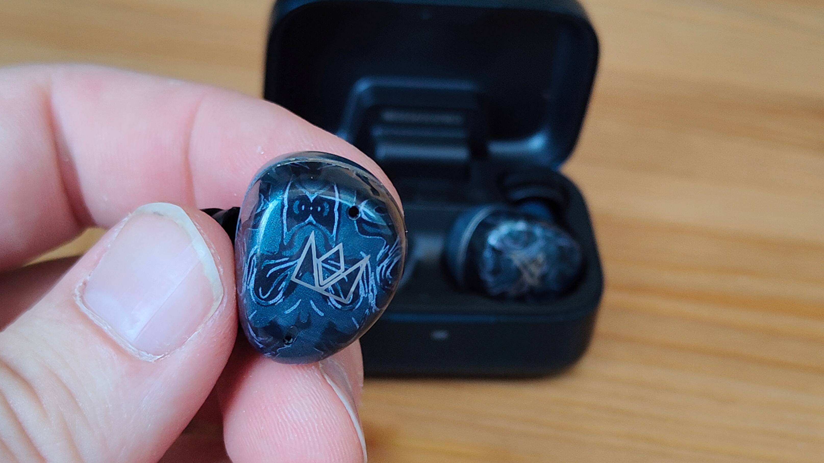 The Noble Osprey earbud in a man's hand, next to the case on a table.