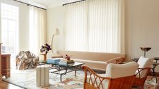 modern living room with beige walls and slightly sheer curtains, a long beige sofa, with two bamboo and boucle armchairs, a wrought iron coffee table, silver sculptural seat, floor lamp, and patterned rug