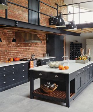 An industrial kitchen with a black and white kitchen island with open shelves, a brick wall behind it with white and black and countertops below it, and a metal walkway above it