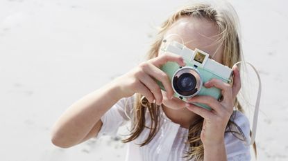 A little blonde girl on a beach taking a picture with one of the best camera for kids