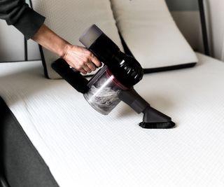 A woman's hand holding a small handheld vacuum cleaner, using an upholstery attachment to vacuum a white mattress.